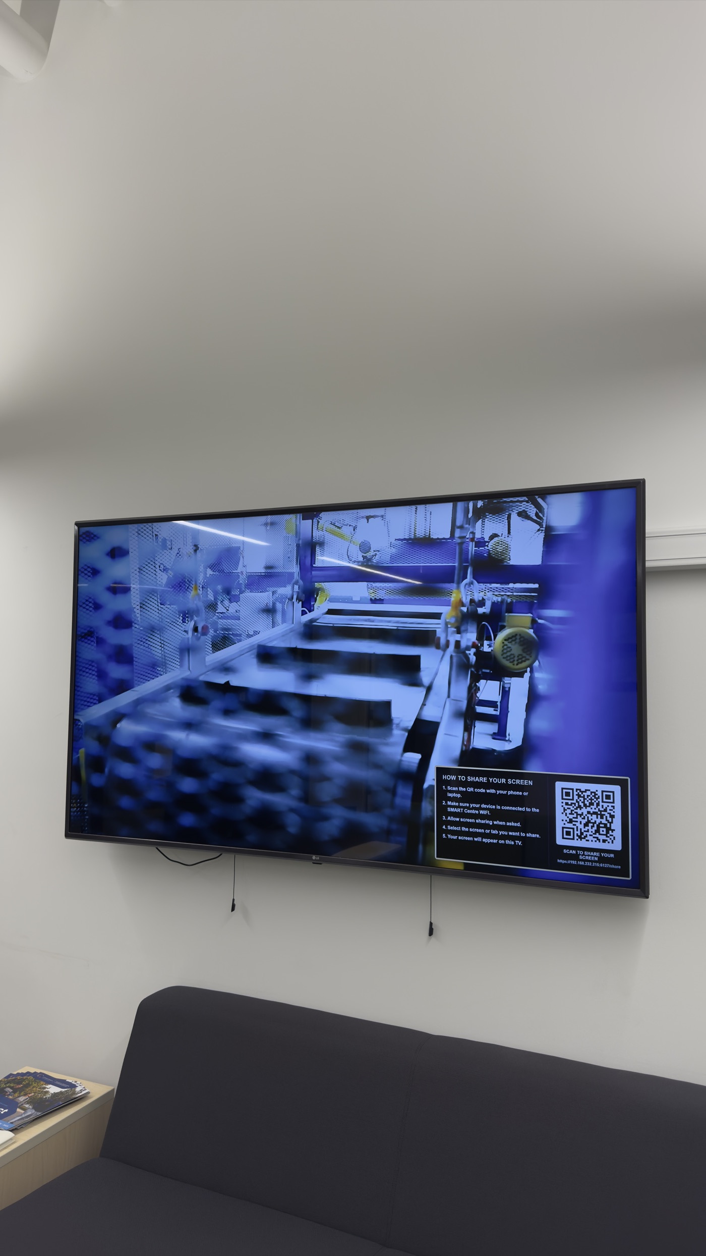 A wall-mounted TV playing a process video with the screen-share overlay and QR code shown in the lower corner