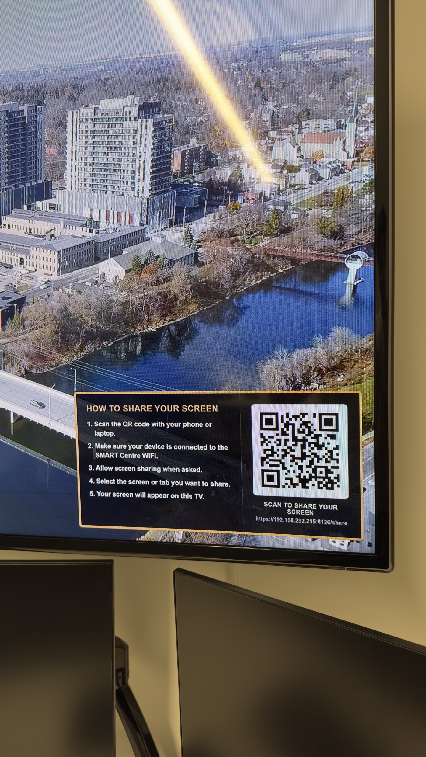 Close-up of a wall-mounted TV showing the screen-share overlay: step-by-step instructions and a QR code labelled Scan to Share Your Screen