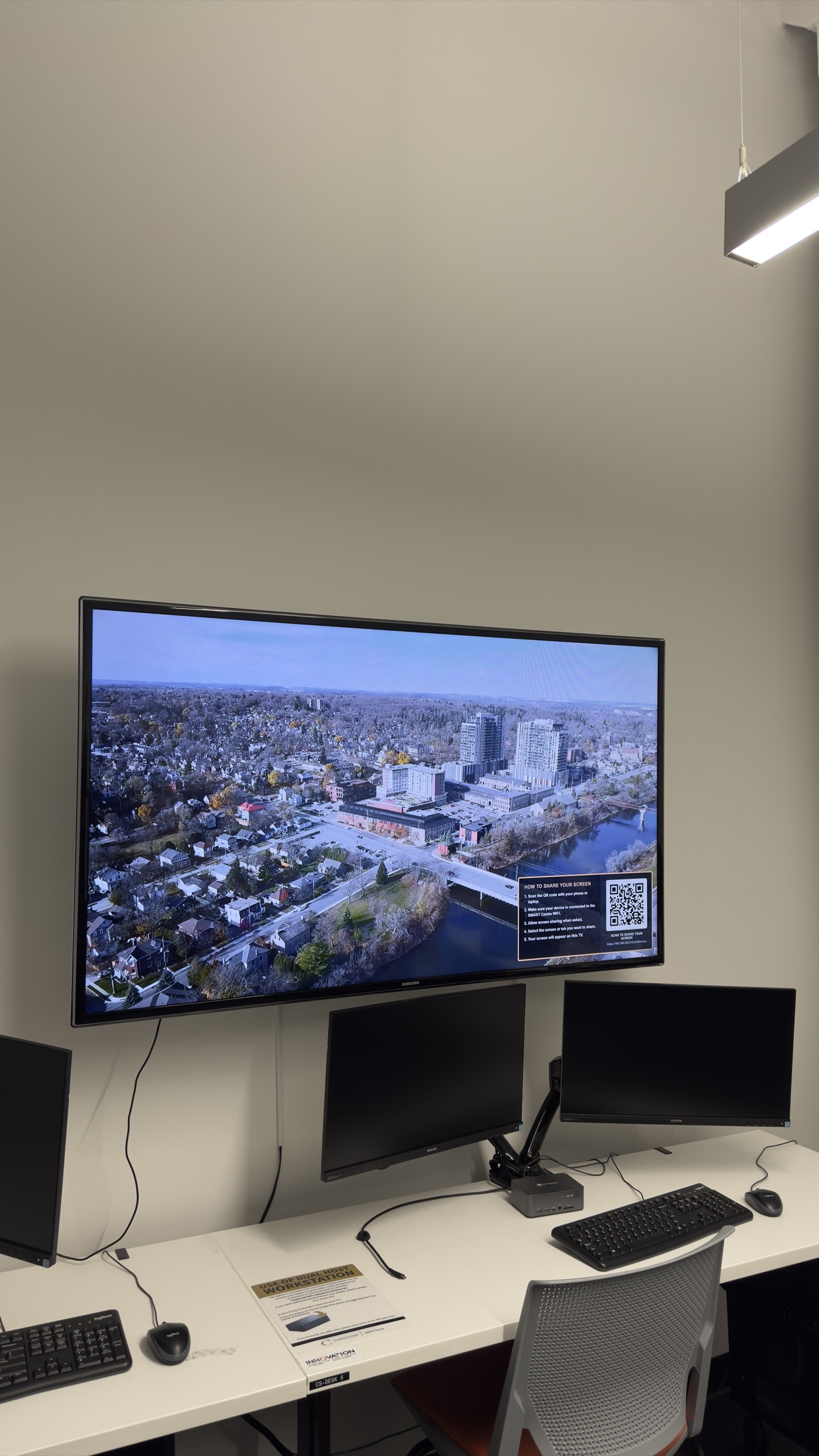 A wall-mounted TV in a workstation area displaying an aerial view of the city with the screen-share overlay in the corner, above a desk with monitors and a mini PC